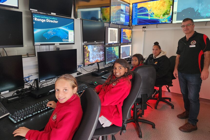 Four students seated in control room with TV screens, teacher standing on right