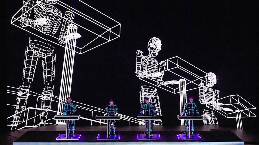 four members of Kraftwerk perform live on stage, lined up at their synths, 3d wire images of robots projected behind