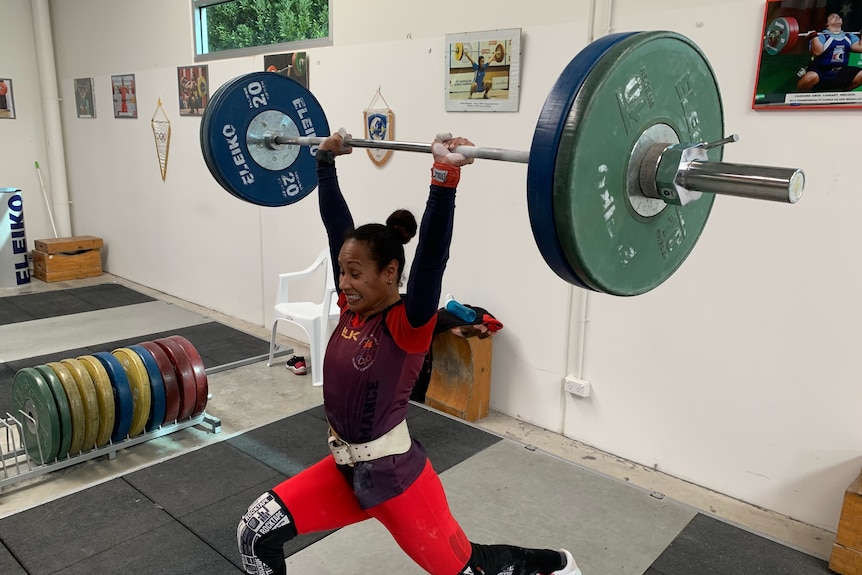 Woman in red unitard lifts barbell and wights over her head while in a lunge.