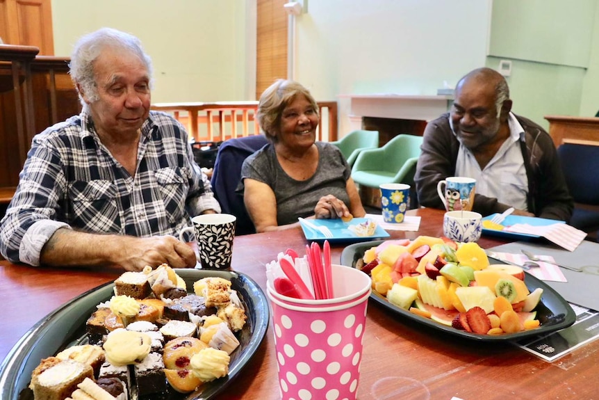 Three people sitting at a table, with plates of cake and fruit on it.
