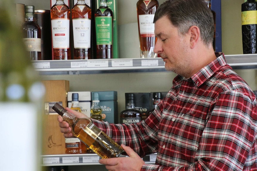 A side-on shot of a man standing in front of shelves with whisky bottles on them, holding one himself and looking at it.