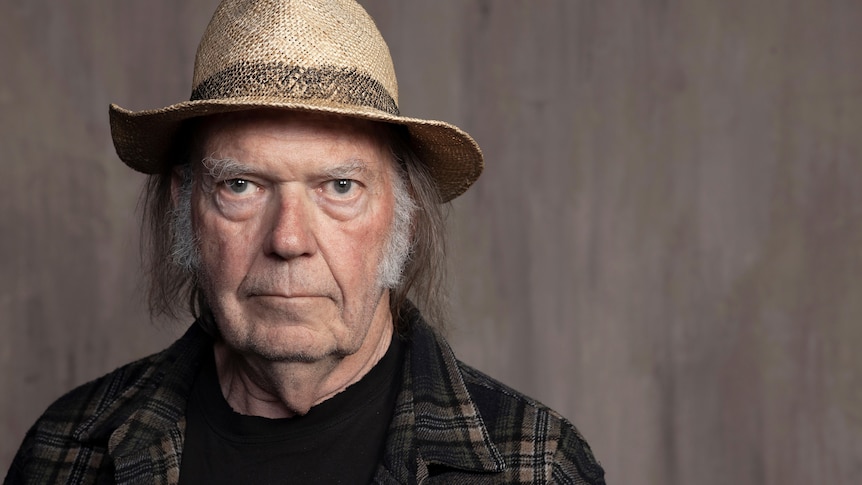 Headshot of Neil Young looking at the camera wearing a straw broad-brimmed hat and checked shirt