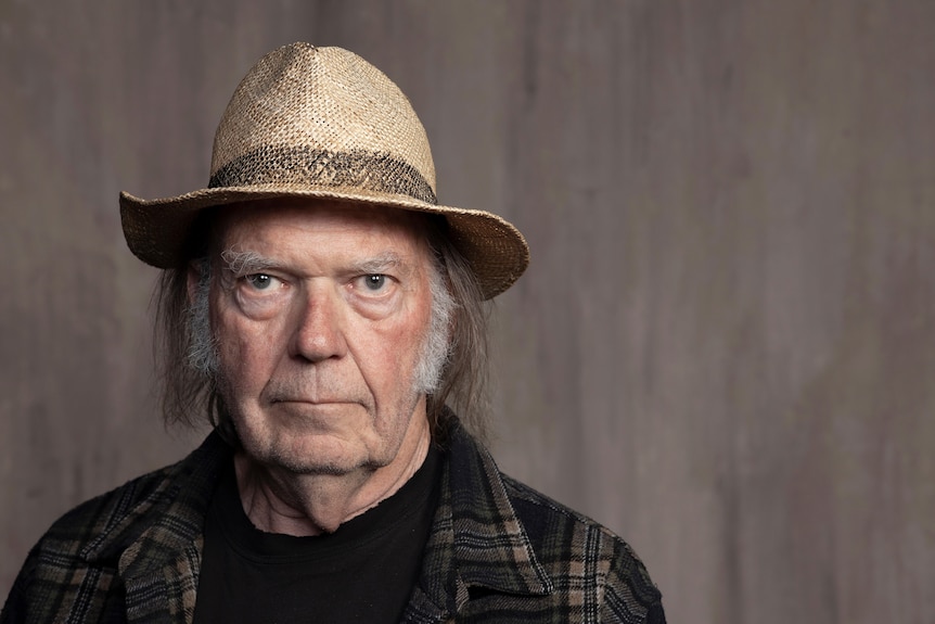 Headshot of Neil Young looking at the camera wearing a straw broad-brimmed hat and checked shirt