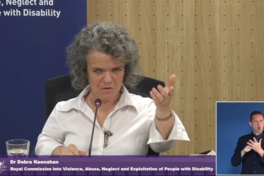 A woman of short stature with grey hair and a white shirt speaking at the royal commission