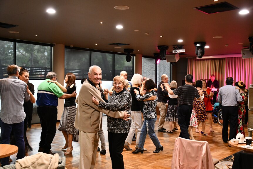 An elderly couple smile and dance in a hall.