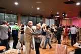 An elderly couple smile and dance in a hall.
