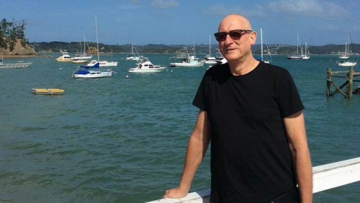 Daryl Oehm wearing a black t-shirt and sunglasses, standing on a pier with boats in background.