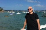 Daryl Oehm wearing a black t-shirt and sunglasses, standing on a pier with boats in background.
