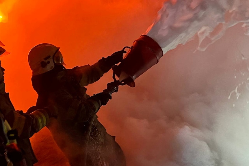 A firefighters throws a bucket of water into smoke and flames.