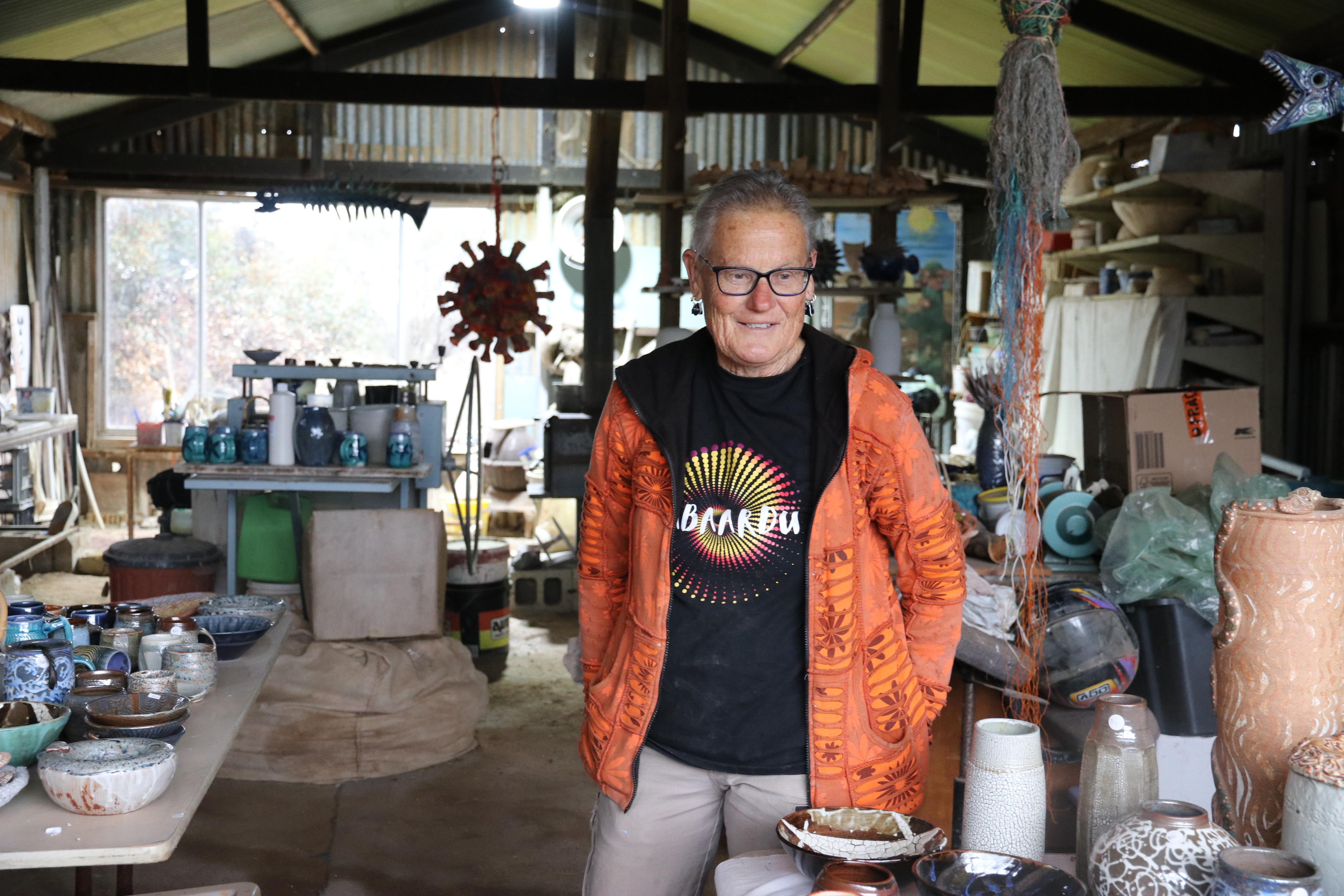 older woman in orange jacket stands in a shed full of pottery 