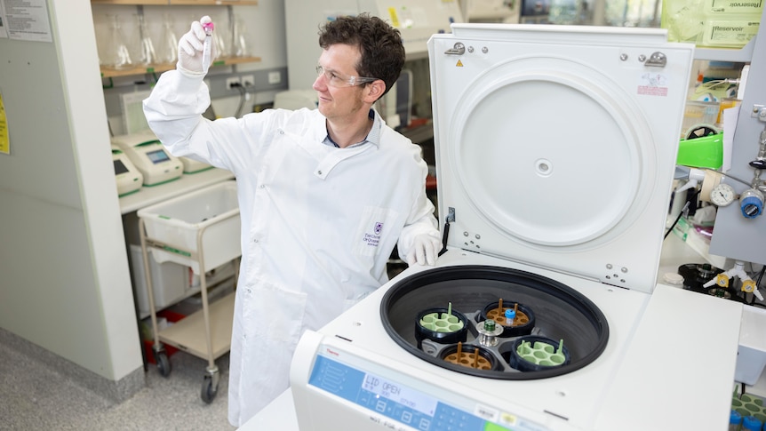 Keith Chappell wearing a lab coat and  holding up a vile next to a large piec of scientific technology. 