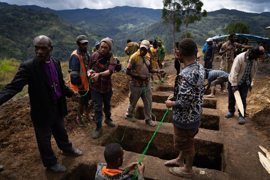 A group of people and a priest standing around graves dug into the dirt