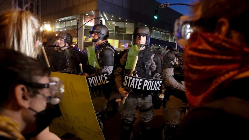 Police wearing combat helmets while carrying shields and large wooden truncheons block the path of a group of civilians.