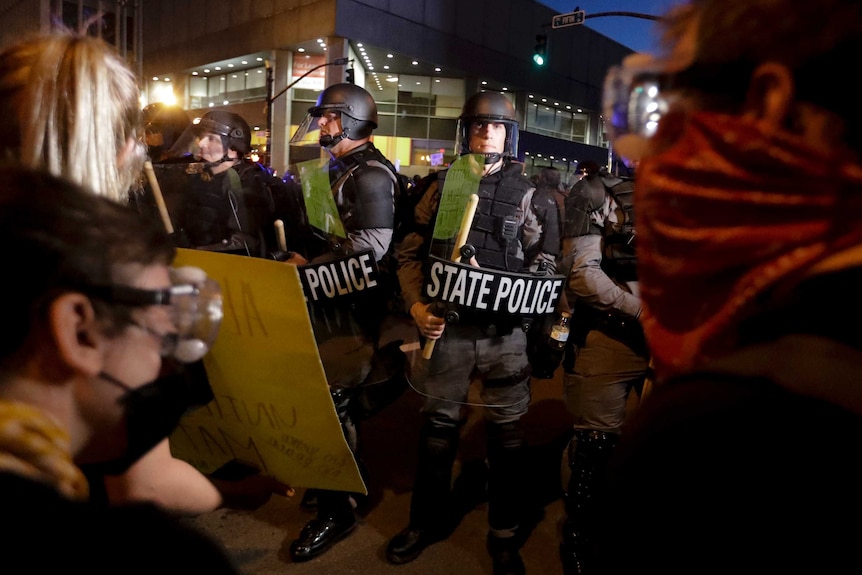 Police wearing combat helmets while carrying shields and large wooden truncheons block the path of a group of civilians.