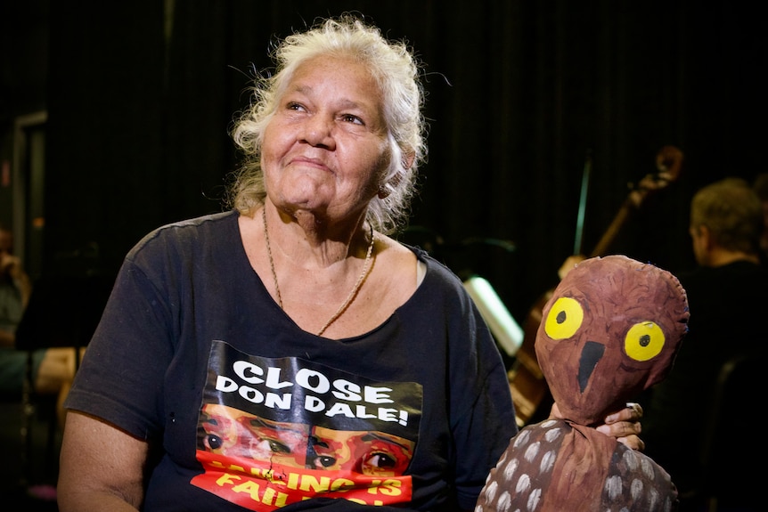 A woman sitting on a darkened stage looking up and smiling, while holding an owl puppet.