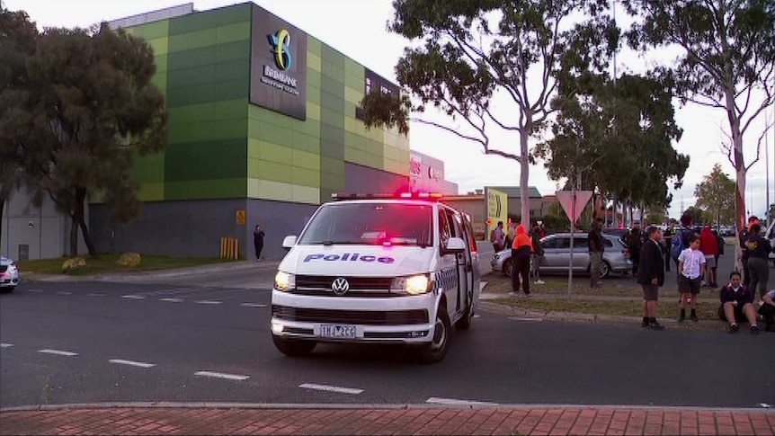 A police van is parked outside a shopping centre with a number of people nearby including children sitting on a curb.