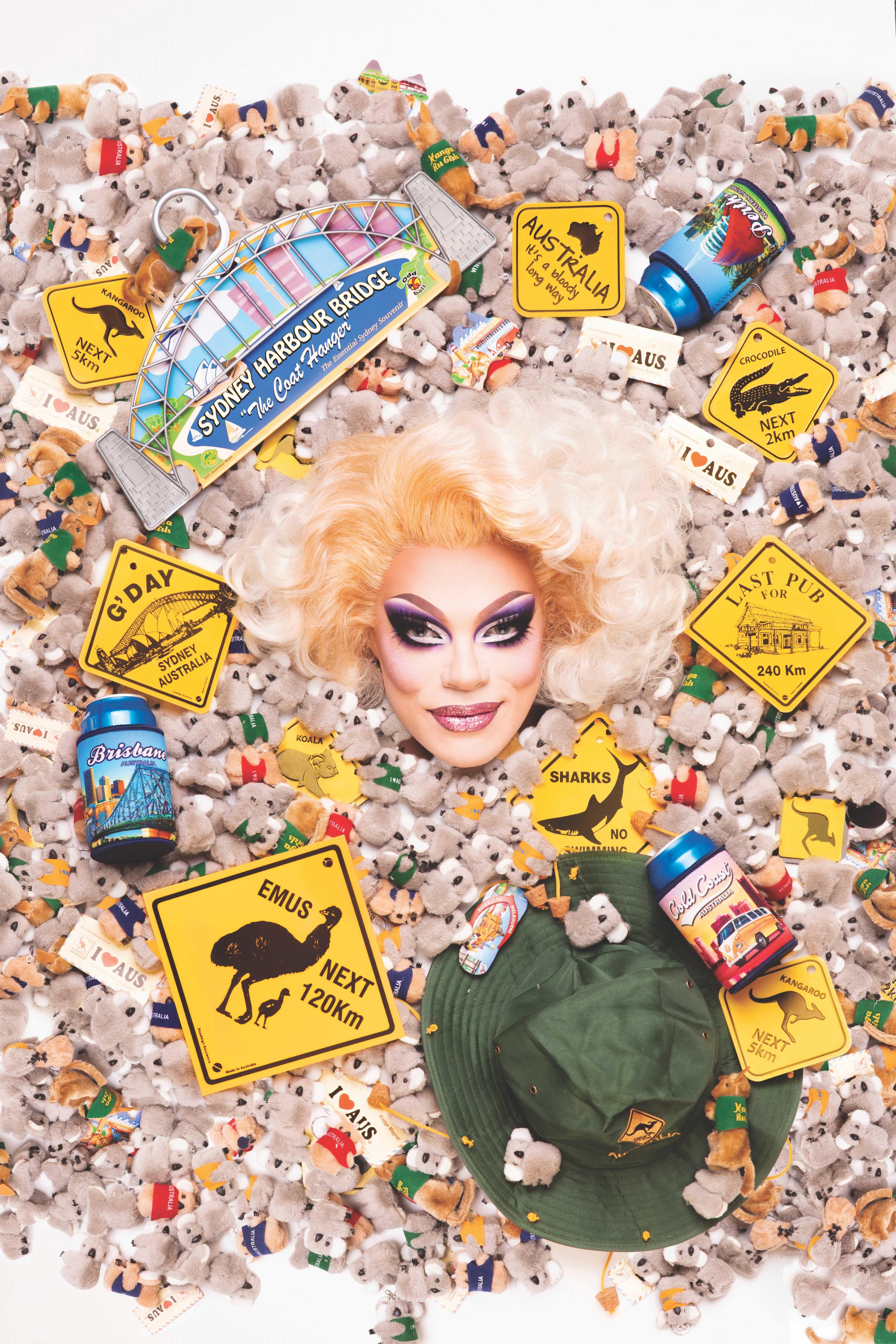 Art Simone in drag makeup and a blonde wig, surrounded by Australiana items including toy koalas and yellow signs that say Gday.