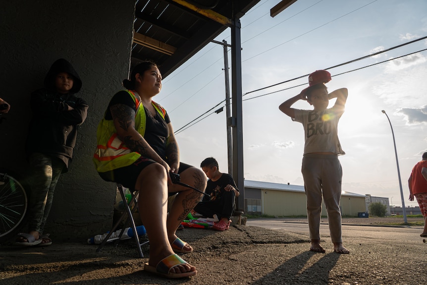 A young woman in a hi vis vest sits at the entrance to a concrete motel driveway. Kids stand around her in the sun.