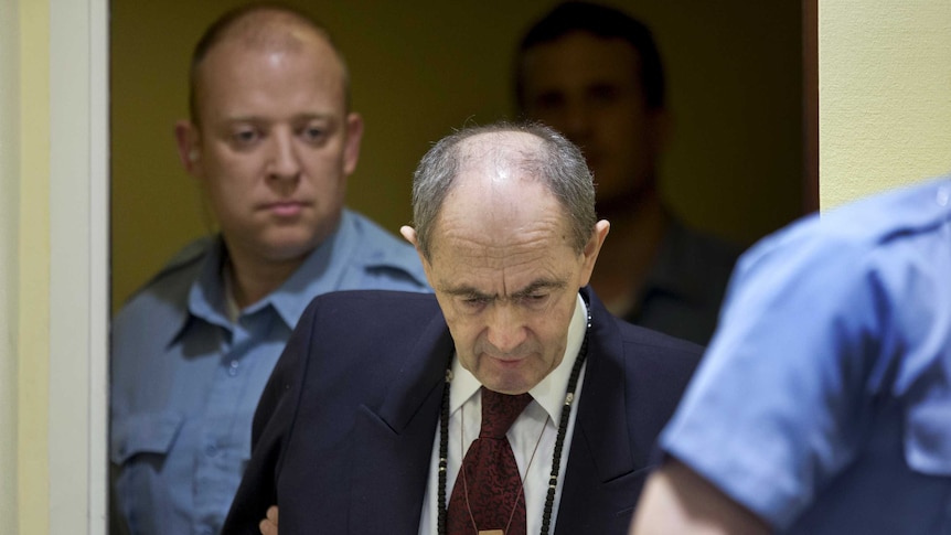 Zdravko Tolimir is escorted by UN security guards in a courtroom.