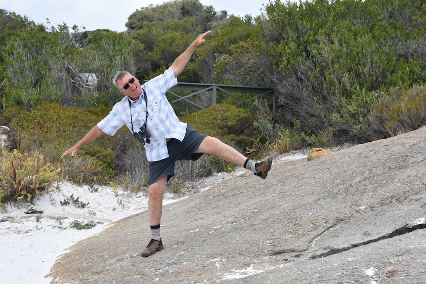 Randall has his arms in the air and a leg up, playfully, on a sloping rock with a camera around his neck.