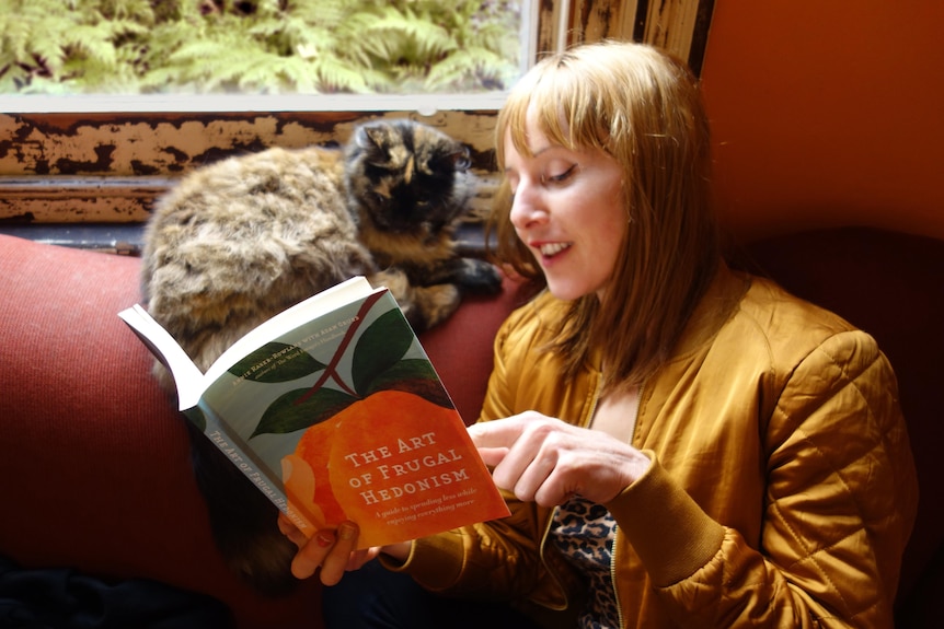 Young woman with ginger hair and fringe appearing to read The Art of Frugal Hedonism on a couch with a tortershell cat