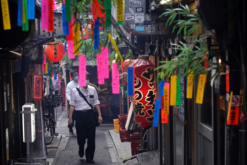 Men wearing face masks walk through a thin alley where many colourful strips of material have been hung outside bars.