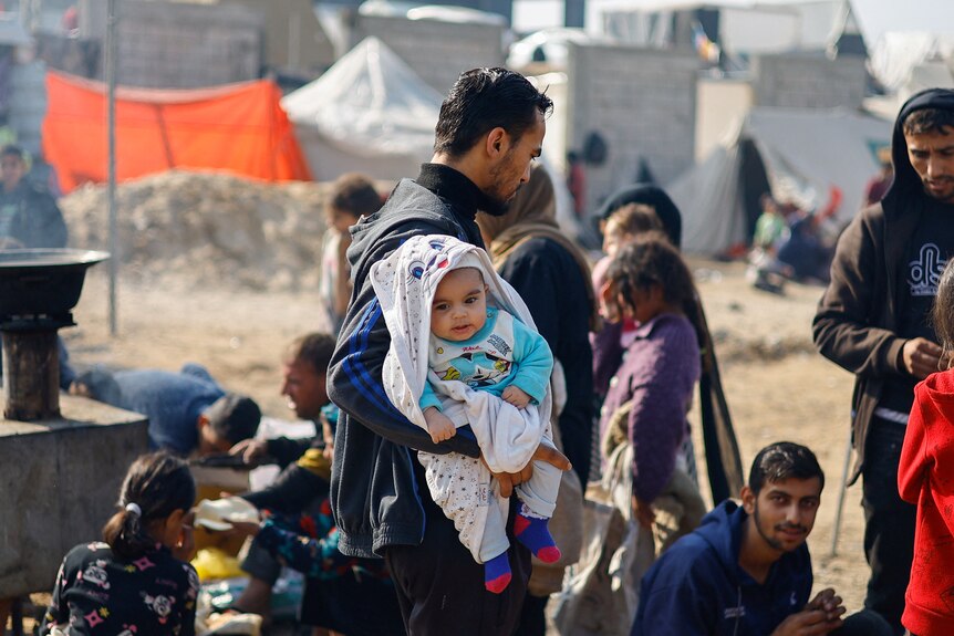 A man looking away holds a baby wearing a blanket 