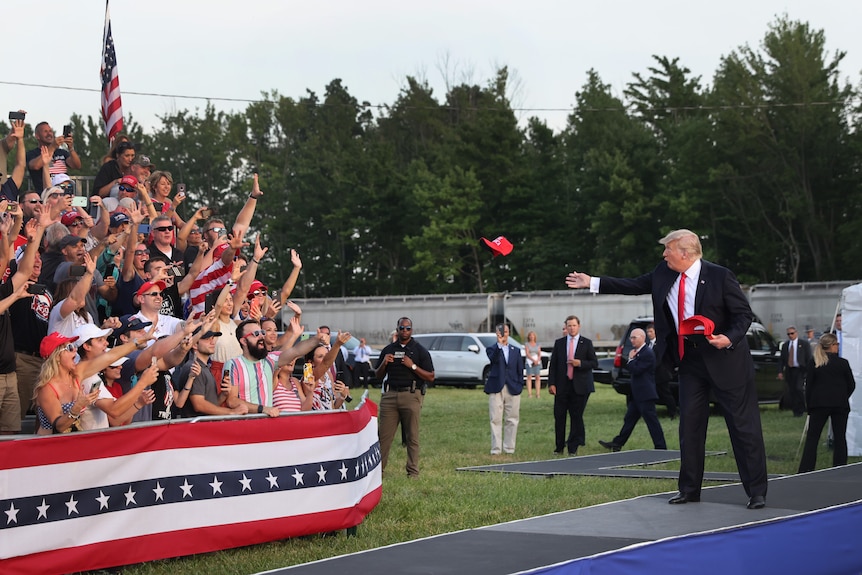 A man in a suit with red tie throws a red cap at enthusiastic crowd