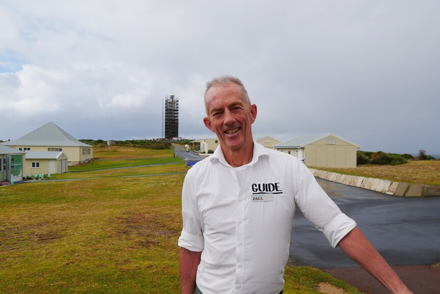 An older man stands in front of a lighthouse. 