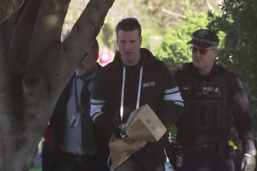 A man wearing paper bags over handcuffs is escorted by two male police officers