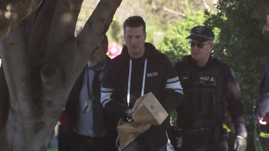 A man wearing paper bags over handcuffs is escorted by two male police officers