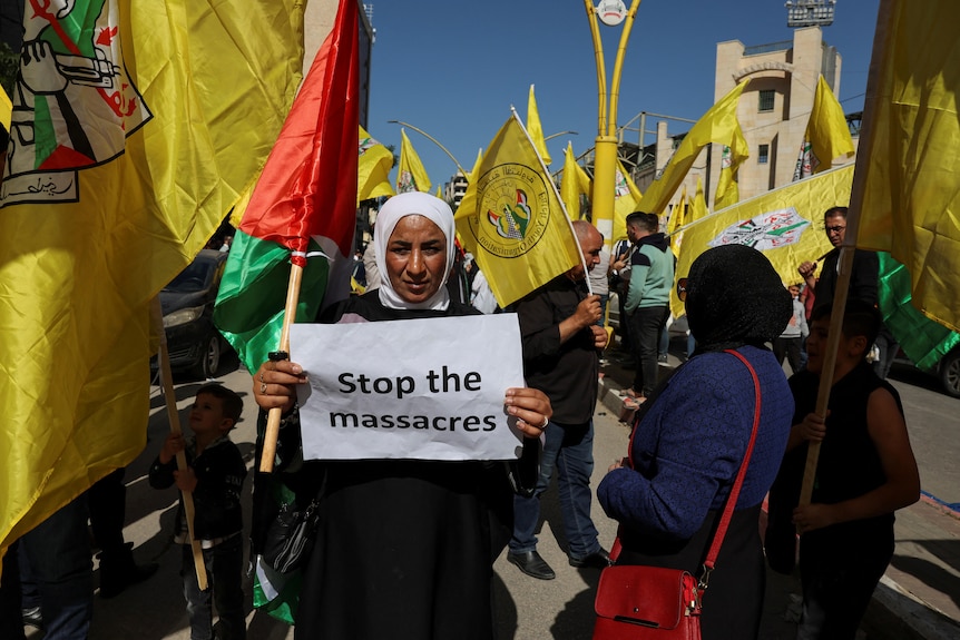 woman holding up a sign with english text "stop the massacres" as well as a palestinian flag