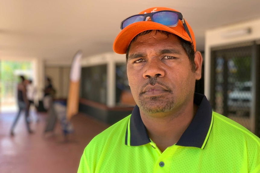 Head and shoulders image of a man in a high-vis shirt and baseball cap standing on sidewalk out the front of a shopping centre.