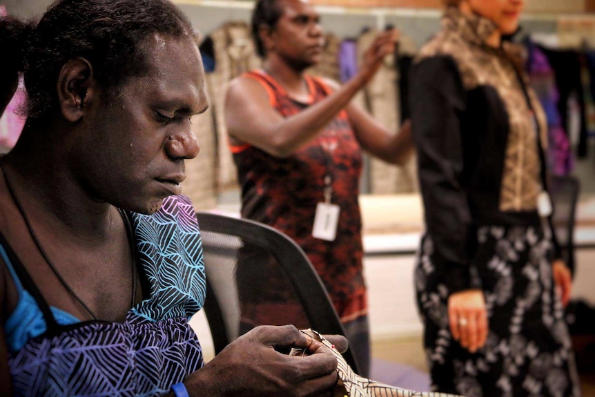 Jamie Timaepatua uses a needle on fabric with a pandanus print while another Sistagirl fits a dress on a model.