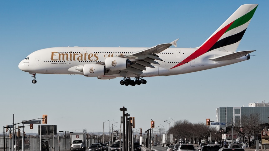 Against a blue sky, a large Emirates A380 lands close to a road, making it appear giant in relation to traffic.