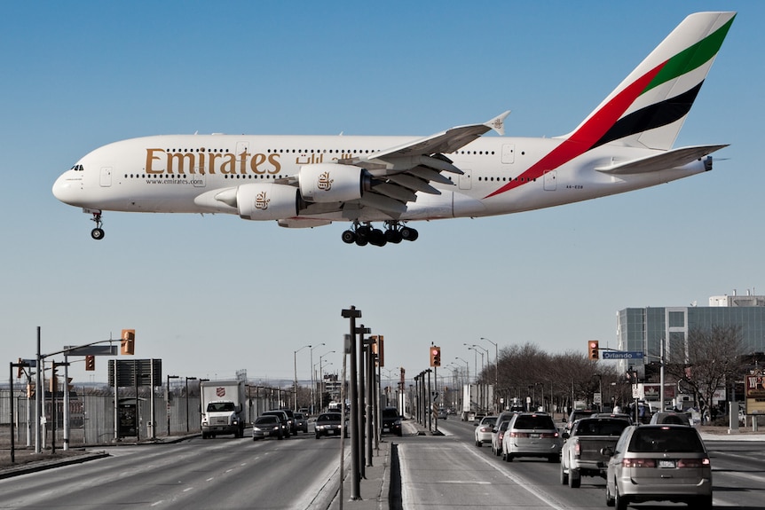 Against a blue sky, a large Emirates A380 lands close to a road, making it appear giant in relation to traffic.