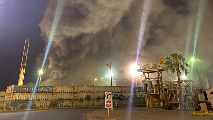 Grey smoke billows into a night sky, behind a stack of cargo.