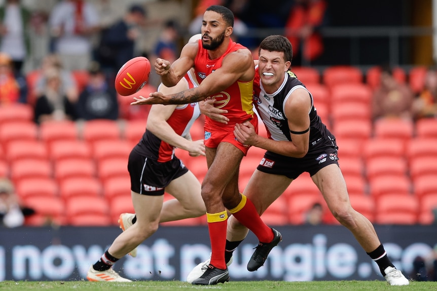 A Gold Coast Suns AFL player handballs while being tackled by a St Kilda opponent.