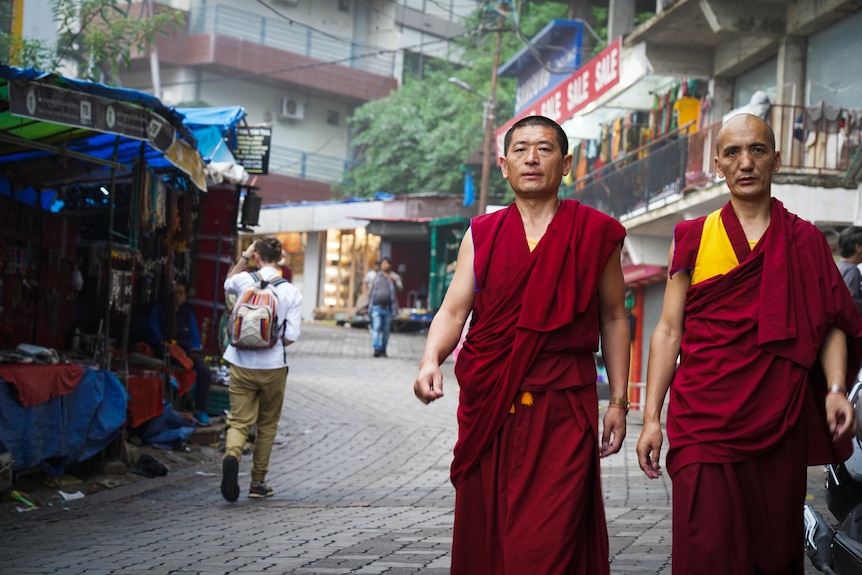 Two monks in traditional maroon and orange vibes walk down a street past stalls and other people
