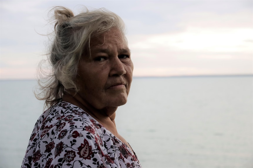 An elderly Indigenous woman standing at a beach with a sunset, looking back at the camera