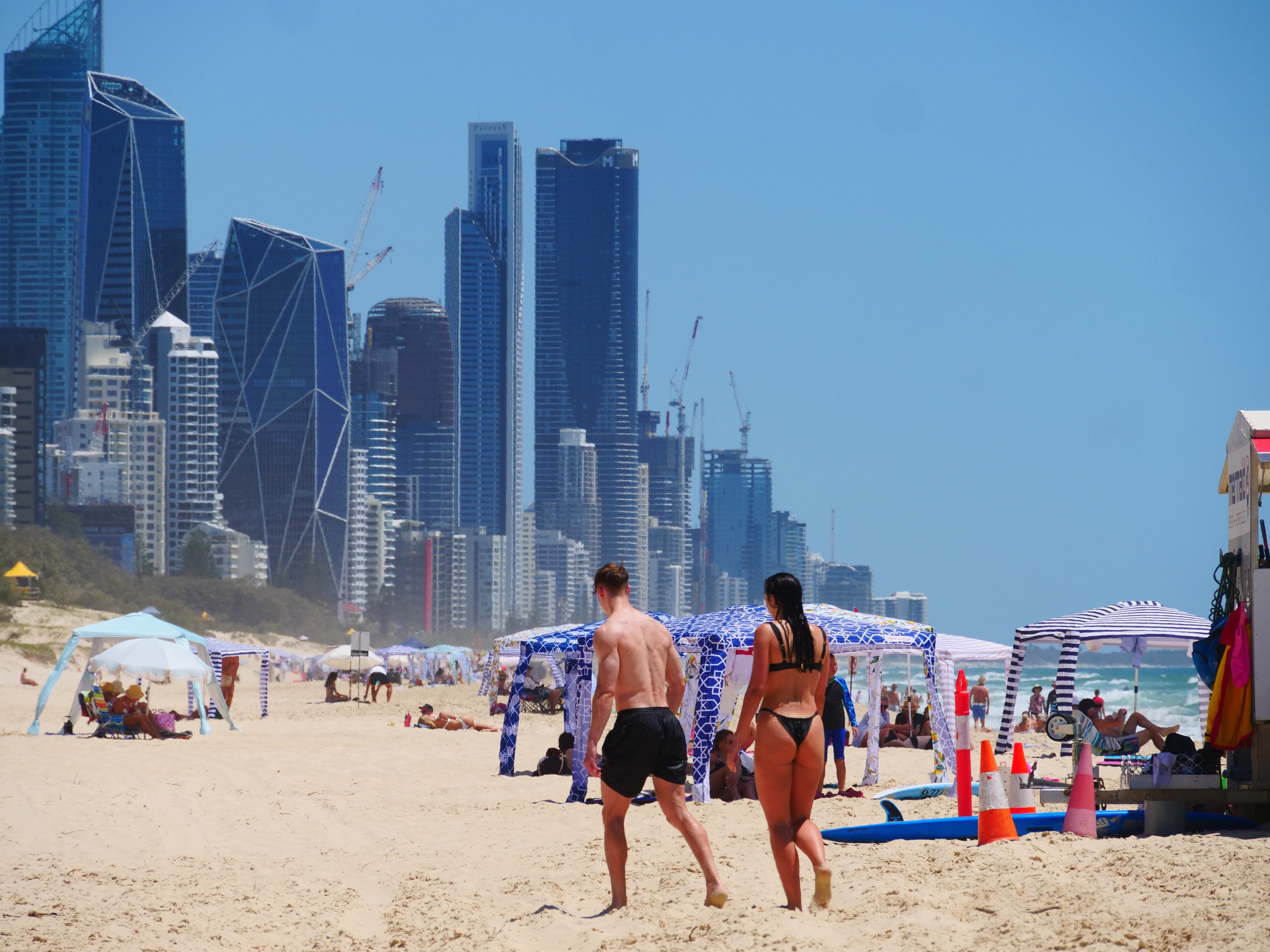 Um homem e uma mulher se afastam da câmera em uma praia lotada próxima ao horizonte da cidade.