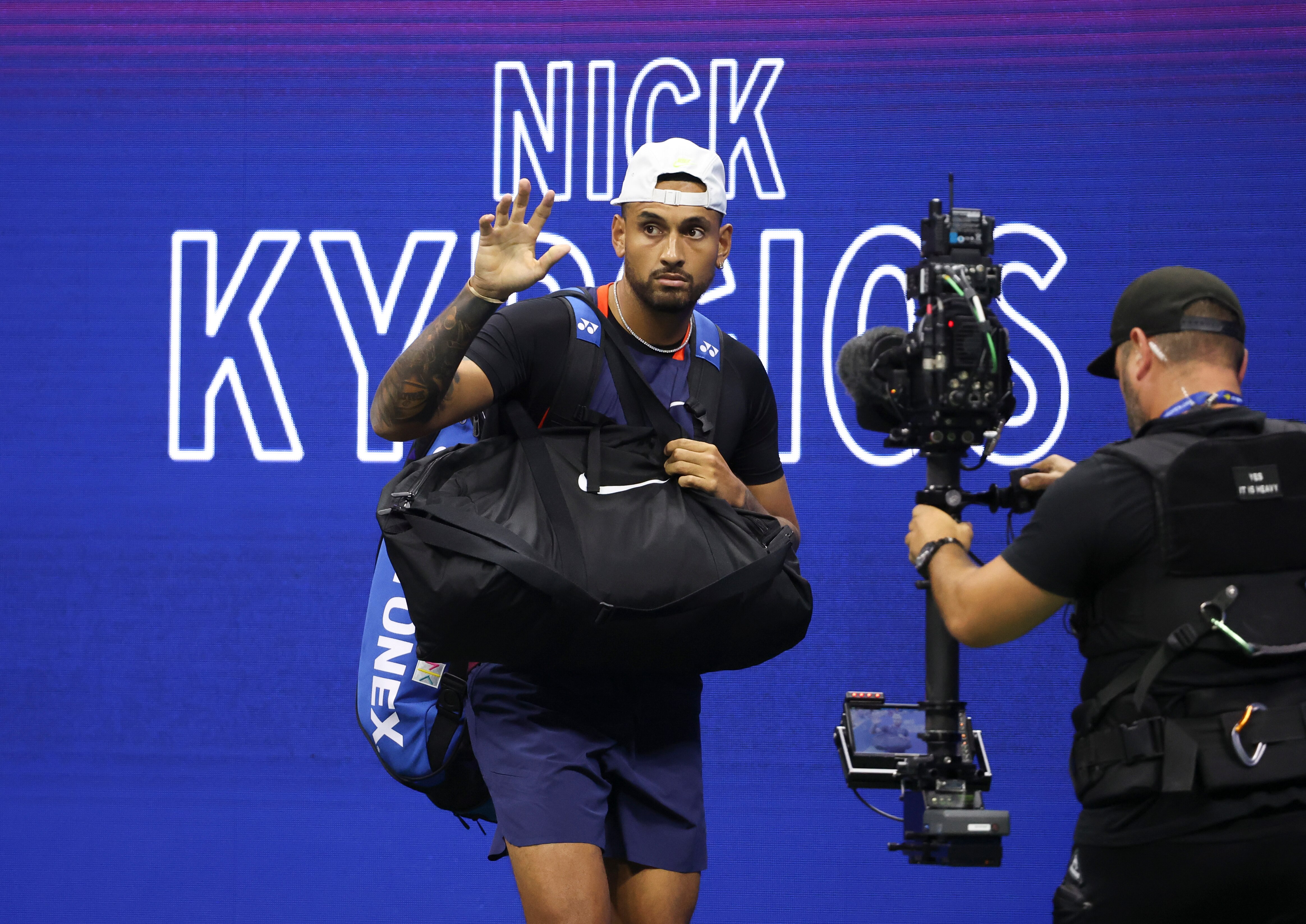 Australian tennis player Nick Kyrgios waves at the crowd as he walks in front of an electronic sign with his name on it.