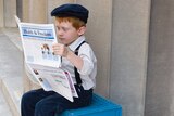 A young boy, dressed in shirt, suspenders and hat, sits on a milk crate reading a newspaper.