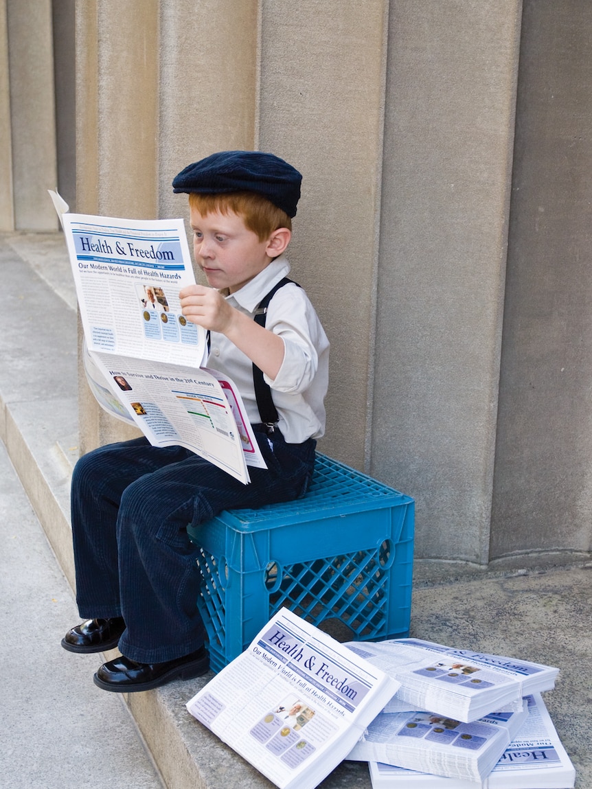 A young boy, dressed in shirt, suspenders and hat, sits on a milk crate reading a newspaper.
