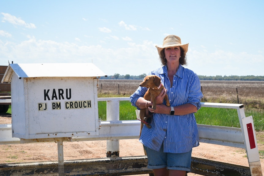 A woman holding a miniature dashound stands next to a cattle grid and white mailbox with the words 'Karu' and 'PJ BM Crouch'.