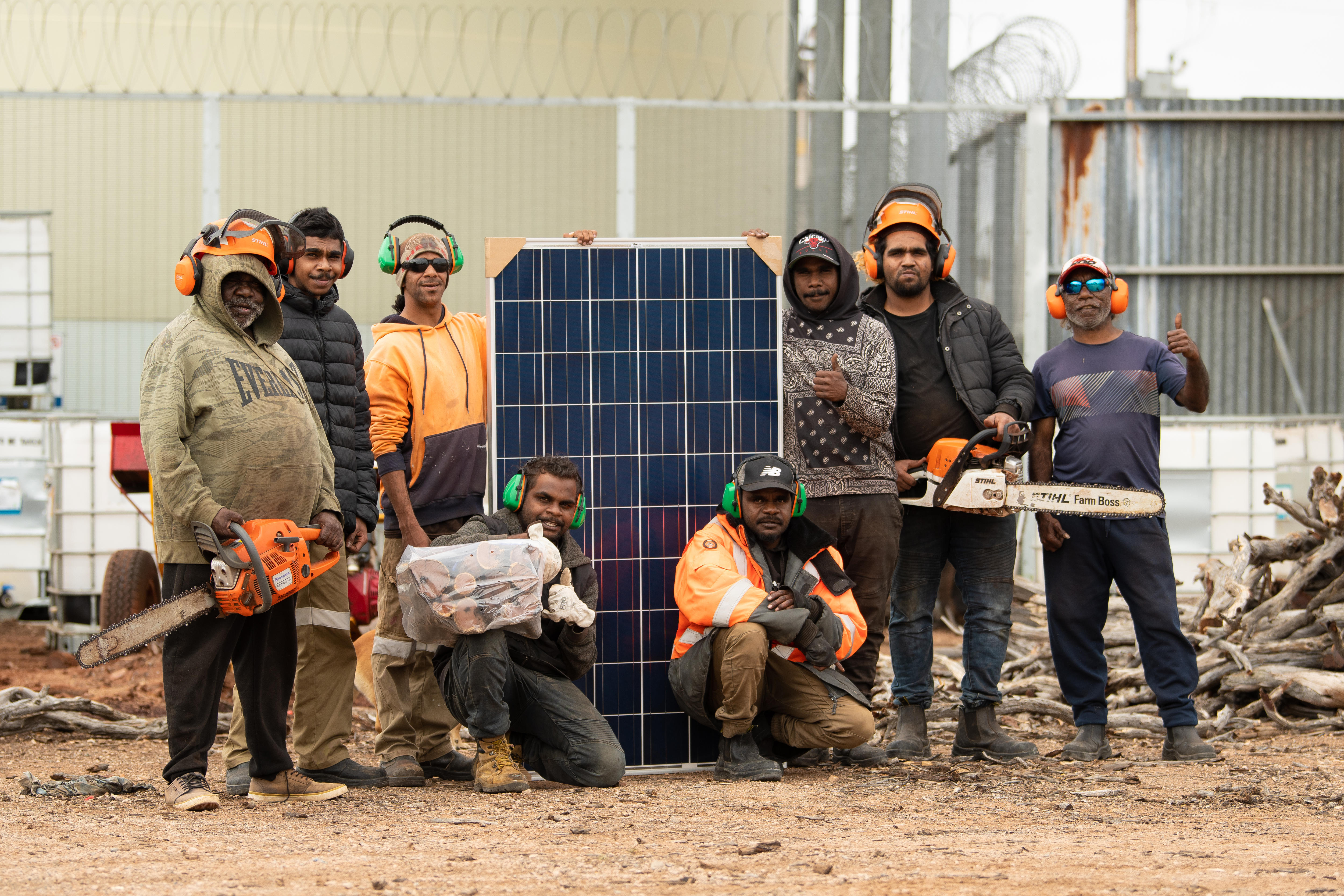 Eight Aboriginal men standing with 1.6 m high solar panel, two holding chainsaws, others with earphones and logs