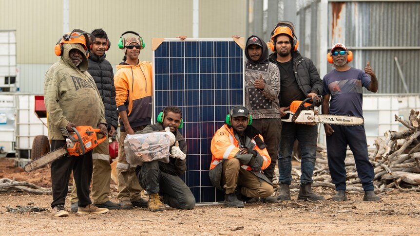 Eight Aboriginal men standing with 1.6 m high solar panel, two holding chainsaws, others with earphones and logs