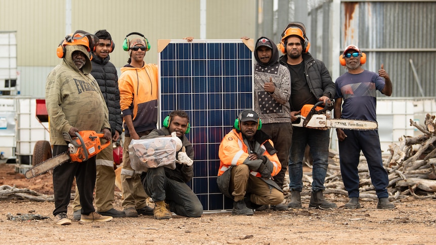 Eight Aboriginal men standing with 1.6 m high solar panel, two holding chainsaws, others with earphones and logs