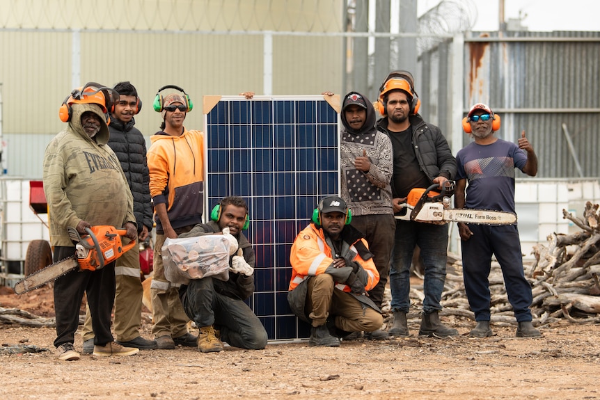 Eight Aboriginal men standing with 1.6 m high solar panel, two holding chainsaws, others with earphones and logs