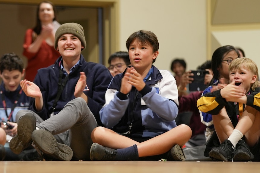 Boys of different ages sit cross-legged on a floor. Some of them are smiling and clapping.